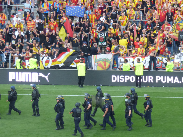 Eine Gruppe von Polizisten in Helmen und mit Waffen in der Hand geht über ein Fußballfeld, im Hintergrund halten Menschen Fahnen und Banner in der Nähe von Geländern.