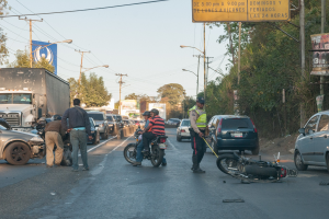 Eine Gruppe von Menschen steht um ein verunglütztes Motorrad auf der Seite einer Straße mit mehreren Fahrzeugen, darunter ein Lastwagen, und einer Hintergrund von Bäumen, Masten, Lichtern und Schildern unter einem Himmel.