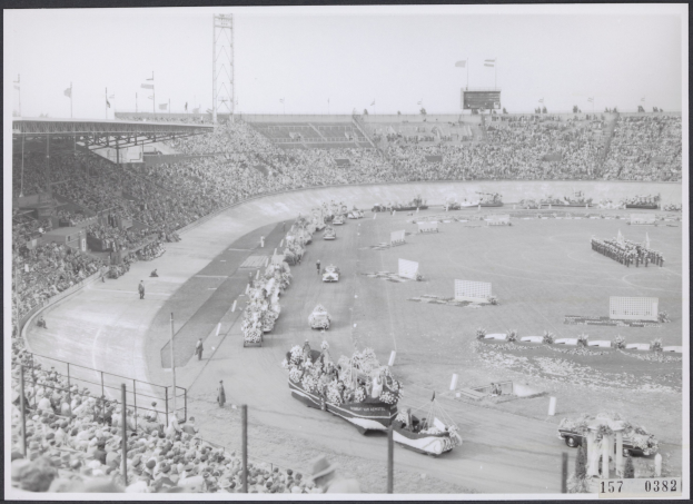 Schwarzes und weißes Foto einer Rennstrecke mit Zuschauern in einem Stadion, einige Fahrzeuge auf der Straße und Pfosten mit Flaggen im Hintergrund.