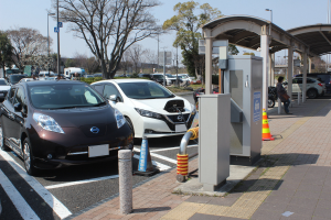 Ladestation für Elektroauto in Japan mit Autos auf der Straße, Verkehrskegeln, einer Person auf dem Gehweg, einem Schuppen, Mästen, Lichtern, Schildern, Bäumen, Pflanzen und einem Himmel im Hintergrund.