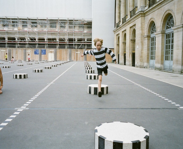 Spielbereich mit vier Kindern; ein Kind läuft auf einer schwarz-weißen Betonstufe in der Nähe eines Schulgebäudes.
