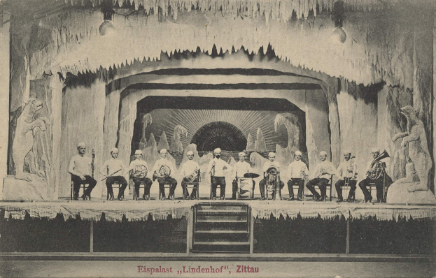 Schwarzes und weißes Foto einer Gruppe von Menschen auf einer Bühne mit Musikinstrumenten, Treppen nach oben, Skulpturen auf beiden Seiten und Text "Lindenhof, Zittau, Deutschland" unten.