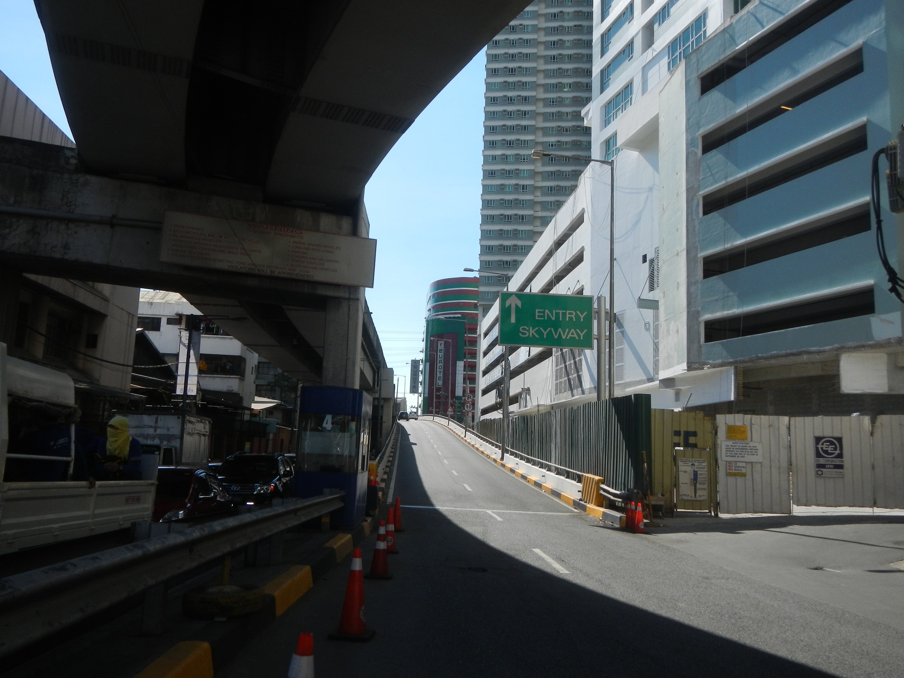 Stadtstraße mit Absperrkegeln, Fahrzeugen, einer Brücke auf der linken Seite, Gebäuden mit Namensschildern, Laternenpfählen und einem Himmel im Hintergrund.