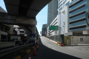 Stadtstraße mit Absperrkegeln, Fahrzeugen, einer Brücke auf der linken Seite, Gebäuden mit Namensschildern, Laternenpfählen und einem Himmel im Hintergrund.