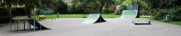 Ein Park mit einer zentralen Skateboard-Rampe, umgeben von Bänken, Gelöndern, Gras, Pflanzen, Bäumen und Gebäuden im Hintergrund.