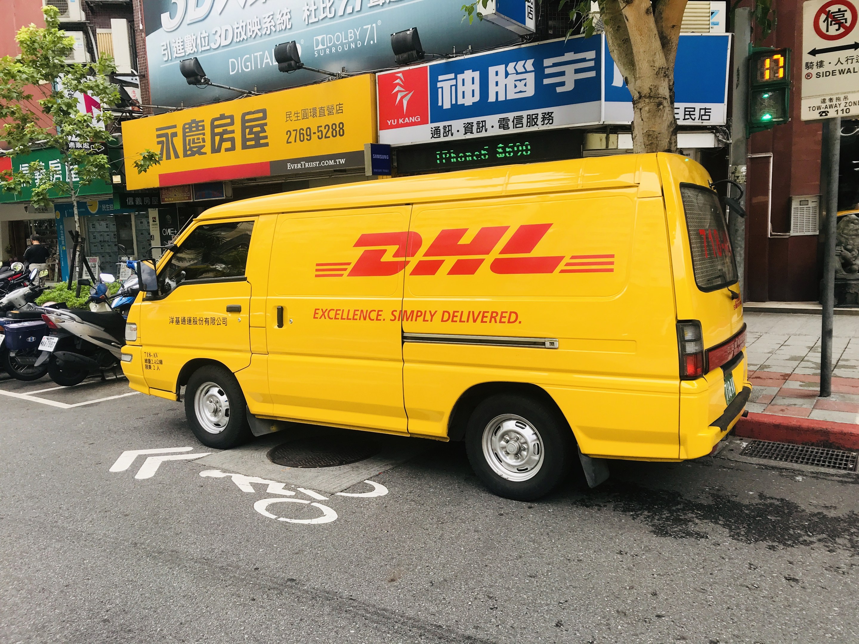 Gelbes DHL-Lieferfahrzeug auf der Straßenseite geparkt, umgeben von Gebäuden, Bäumen, Schildern, Strommästen, Ampeln und einem Fussgänger auf dem Gehweg.