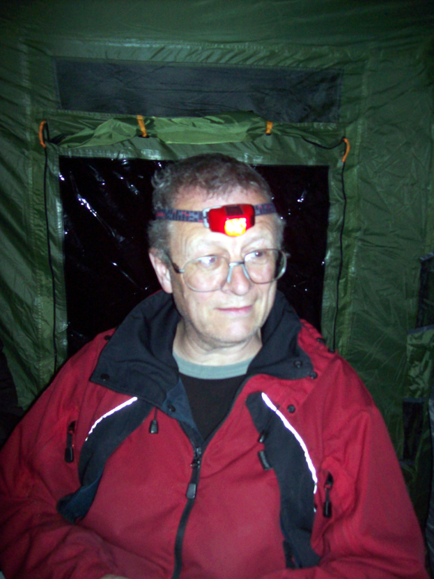 Ein alter Mann in einer roten Jacke ist in einem Zelt und trägt eine Stirnlampen.