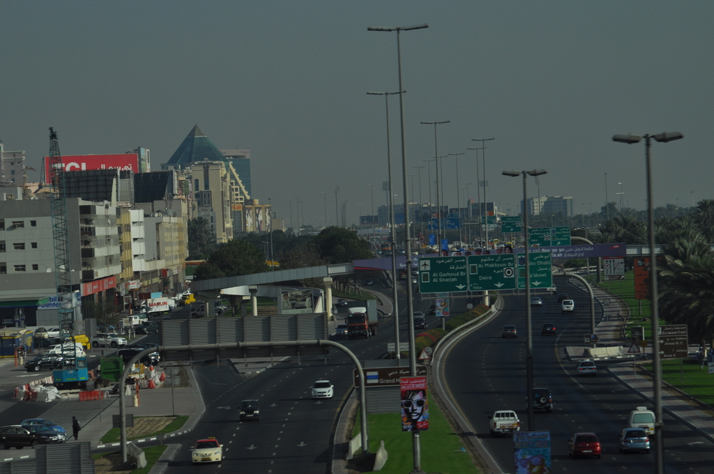 Eine Luftaufnahme einer Stadt mit Gebäuden, Türmen, Fahrzeugen, Werbetafeln, Schildern, Straßen, Bäumen, Straßenlaternen, Menschen, Verkehrszeichen und Informationsanzeigen unter einem klaren Himmel.
