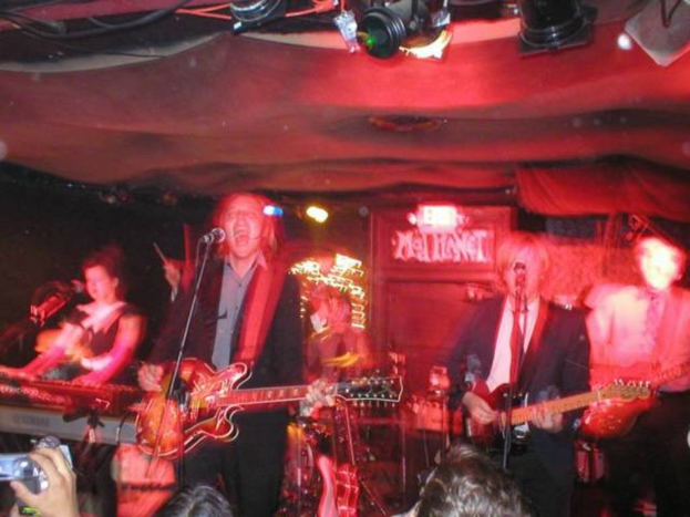 Eine Gruppe von Menschen singt auf der Bühne mit Gitarren, während andere vorne die Performance mit ihren Geräten aufnehmen.