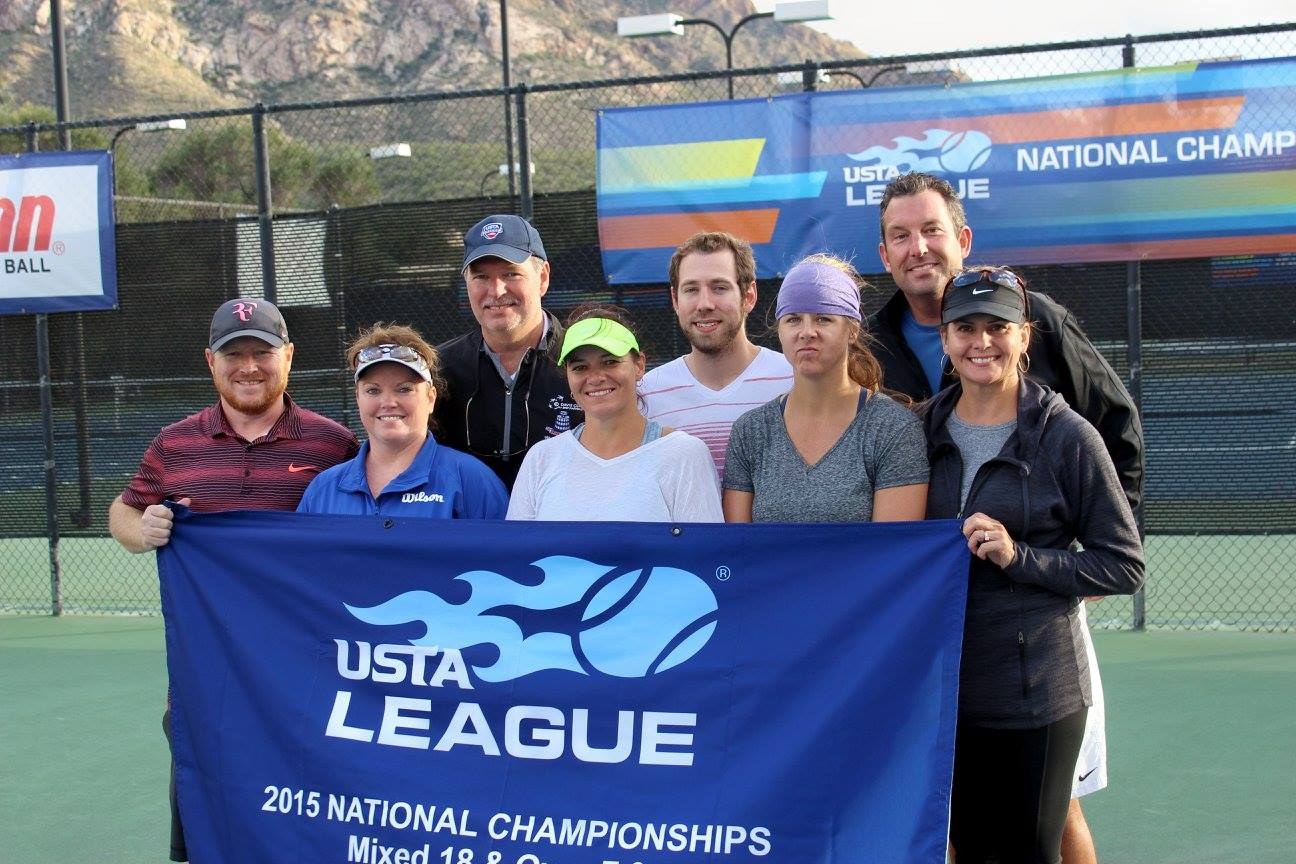 Gruppe von Menschen auf einem Tennisplatz mit einem "2015 USTA National Championships"-Schild, einem Zaun, Bannern, Laternenmasten, Bäumen, Bergen und einem klaren blauen Himmel im Hintergrund.