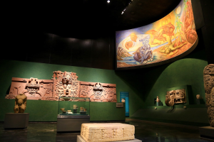 Ein Museumsraum mit verschiedenen Artefakten, darunter Skulpturen, Tischobjekte, eine Texttafel, ein Wandgemälde und Deckenbeleuchtung im Mexiko-Stadt-Museum der Anthropologie.