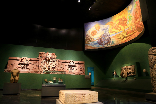 Ein Museumsraum mit verschiedenen Artefakten, darunter Skulpturen, Tischobjekte, eine Texttafel, ein Wandgemälde und Deckenbeleuchtung im Mexiko-Stadt-Museum der Anthropologie.