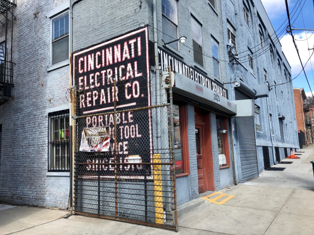 Außenansicht des Cincinnati Electrical Repair Co. Gebäudes mit einer Schautafel, Metallzaun, Mülltonne, Pfählen, Drähten, Bäumen und einem bewölkten Himmel.