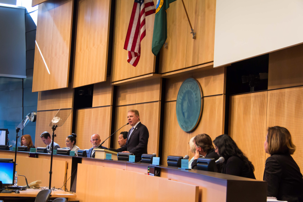 Eine Gruppe von Menschen sitzt an einem Tisch mit Namensschildern, Mikrofonen und anderen Gegenständen, die einem Bildschirm zugewandt sind, mit einem Mann in der Mitte, und Fahnen und einer Tafel im Hintergrund während einer Ratssitzung.