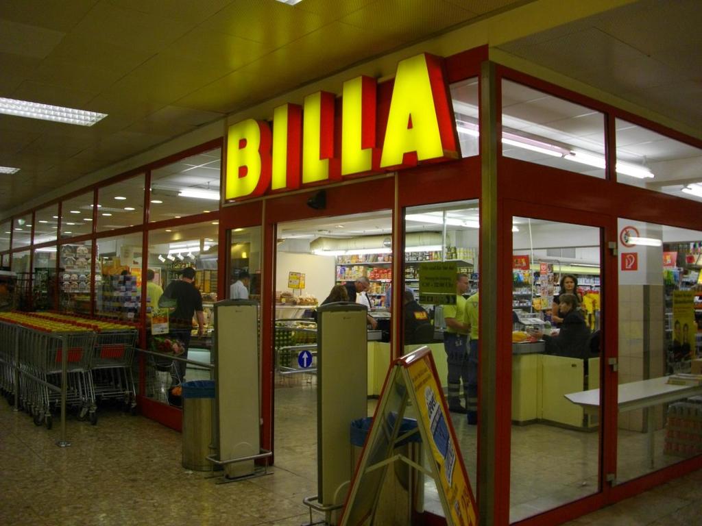 Ein Markt mit einem Eingang in der Mitte, Mülltonnen daneben, einer Reihe von Wagen auf der linken Seite, Deckenbeleuchtung und einigen Menschen, die Produkte durchstöbern.