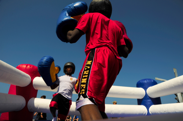 Zwei Boxer in einem Boxring, einer in einem roten Hemd und blauen Shorts, kämpfen mit Boxhandschuhen, umgeben von ein paar Menschen und unter einem klaren blauen Himmel.