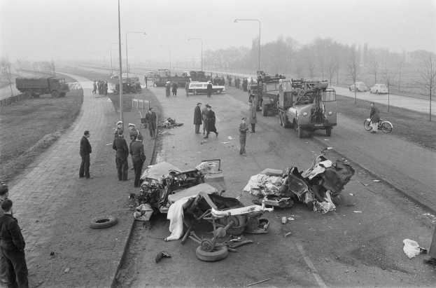 Eine Schwarz-Weiß-Szene eines Autounfalls am Straßenrand mit mehreren Fahrzeugen, darunter ein Landfahrzeug, und einer Gruppe von Menschen drumherum, mit Laternenpfählen, Bäumen und Himmel im Hintergrund.