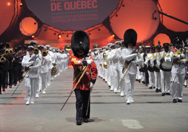 Eine Gruppe uniformierter Personen marschiert auf einer Straße, einige spielen Musikinstrumente, mit einem beleuchteten Schild im Hintergrund, auf dem "Montreal International Festival of Military Bands" steht.