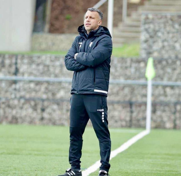 Ein Mann auf einem Fußballfeld, der eine schwarze Jacke und Hose trägt und mit verschränkten Armen und entschlossener Miene in der Mitte eines Trainings steht.