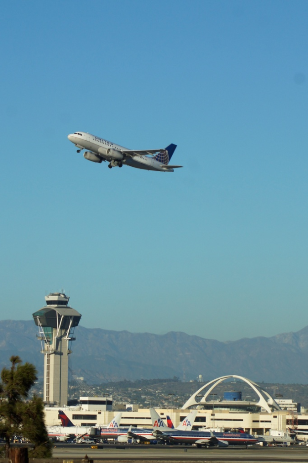 Eine Flughafenansicht mit einem weißen Kontrollturm im Vordergrund, parkenden Flugzeugen am Boden, entfernten Gebäuden und einem Flugzeug am Himmel.