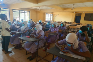 Ein Klassenzimmer in Nigeria mit Schülern, die Gesichtsmasken tragen und auf Bänken oder Stehplätzen sitzen, mit Büchern und Gegenständen auf den Tischen, Fenstern, einer Tür und Deckenleuchten und -ventilatoren im Hintergrund.