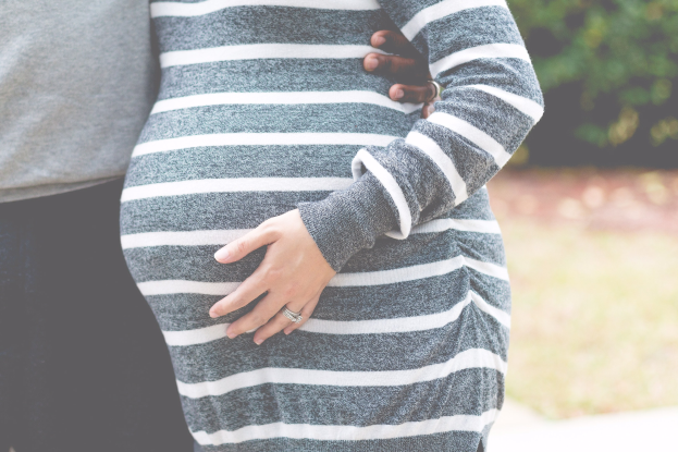 Schwangeres Paar steht zusammen mit den Händen auf dem Bauch, trägt Schwangerschaftskleidung, in einer holzigen Outdoor-Umgebung.