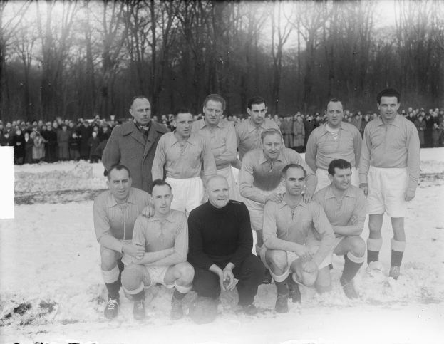 Ein Schwarz-Weiß-Foto einer Fußballmannschaft, die im Schnee posiert, wobei einige Spieler stehen und andere sitzen und im Hintergrund Bäume und ein klarer Himmel zu sehen sind.