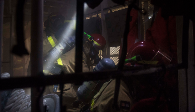 Eine Gruppe von Feuerwehrleuten in Helmen und Schutzausrüstung, einer hält einen Feuerlöscher, mit einem Metallgitter und Seil im Vordergrund und verschiedenen Gegenständen im Hintergrund.