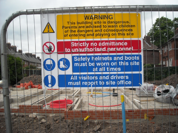 Eine Warnschild ist an einem Tor vor einer Baustelle angebracht, mit Häusern und Bäumen im Hintergrund.