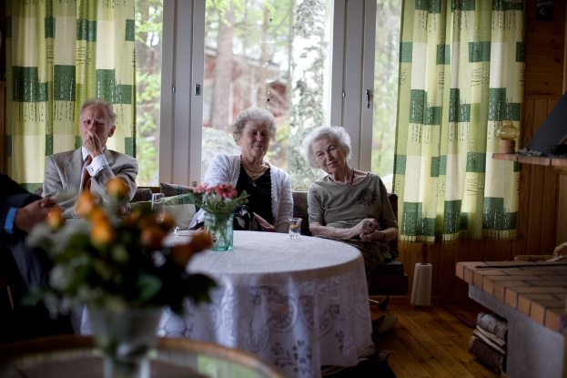 Eine Gruppe älterer Menschen sitzt auf einem Sofa mit einem Tisch davor, hält eine Vase mit Blumen und eine Glastür mit Vorhängen dahinter.