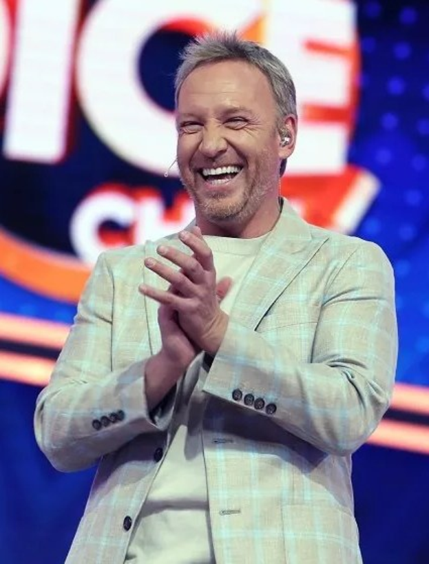 Ein Mann im Anzug klatscht auf einer Bühne mit 'The Voice'-Logo im Hintergrund.