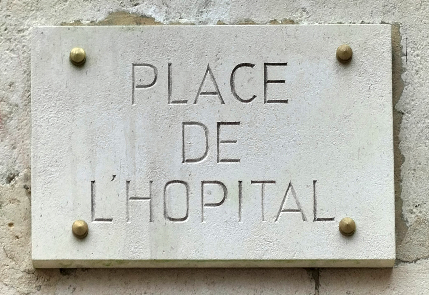 Ein hellgrauer Steinplatten auf einer Gebäudewand trägt die Aufschrift 'Place de l'Hospital' in schwarzer Schrift.