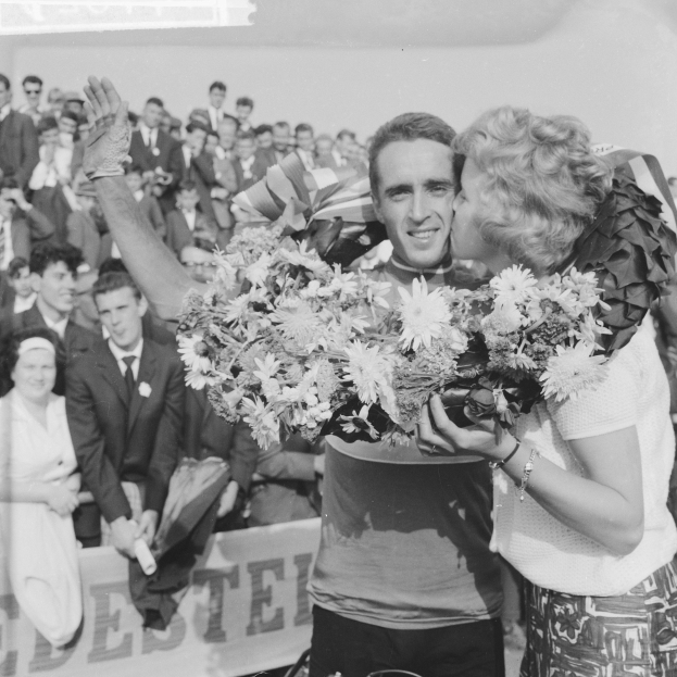 Ein Schwarz-Weiß-Foto eines Mannes und einer Frau, die sich in einer Menschenmenge umarmen und küssen, während der Mann einen Blumenstrauß hält und die Frau strahlt, während die Menge applaudiert.