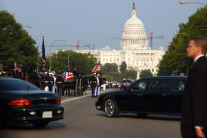 Trauerzug mit Menschen in Mützen und Fahnen vor dem Kapitolsgebäude, Fahrzeuge auf der Straße, Laternenpfähle, Bäume und Gebäude im Hintergrund unter einem klaren Himmel.