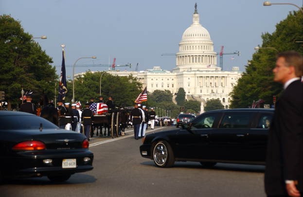 Trauerzug mit Menschen in Mützen und Fahnen vor dem Kapitolsgebäude, Fahrzeuge auf der Straße, Laternenpfähle, Bäume und Gebäude im Hintergrund unter einem klaren Himmel.