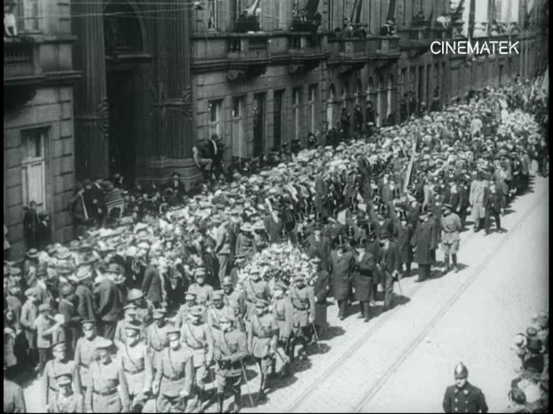 Schwarzes Foto einer großen Menge, die vor einem Gebäude marschiert, einige halten Gewehre, mit einem Wasserzeichen oben rechts.