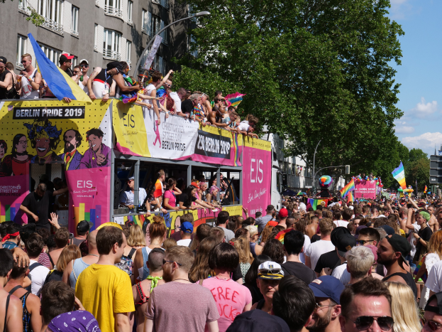 Eine große Menschenmenge marschiert auf der Straße bei der Berlin Pride 2019, viele tragen Mützen und Schutzbrillen, halten Fahnen und Schilder, mit Gebäuden im Hintergrund bei bewölktem Himmel.