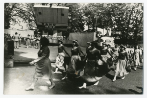 Eine Gruppe von Frauen, die auf der Straße tanzen, umgeben von einer Menge, einem Lautsprecher, einem Fahrzeug, einer Tafel mit Text und einer Fahnenstange.