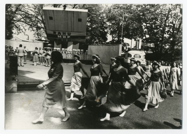 Eine Gruppe von Frauen, die auf der Straße tanzen, umgeben von einer Menge, einem Lautsprecher, einem Fahrzeug, einer Tafel mit Text und einer Fahnenstange.