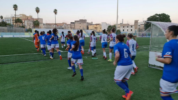 Eine Gruppe von Frauen, die auf einem Feld mit Gebäuden, Bäumen und Pfählen umgeben und einem klaren blauen Himmel Fußball spielen, mit einem Torpfosten und Netz auf der rechten Seite.