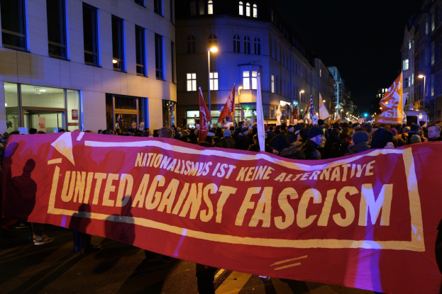Eine Gruppe von Menschen marschiert auf einer Straße, hält ein Banner mit der Aufschrift "Einheit gegen Faschismus", mit Gebäuden, Laternen und Schildern an der Straße und einem sichtbaren Himmel.