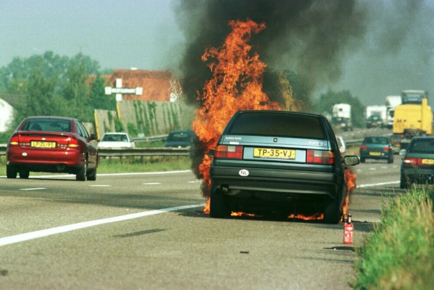 Ein Auto brennt am Straßenrand mit anderen Fahrzeugen in der Nähe, umgeben von Bäumen, Gebäuden und einem klaren blauen Himmel, mit Gras und einem Feuerlöscher auf der rechten Seite.