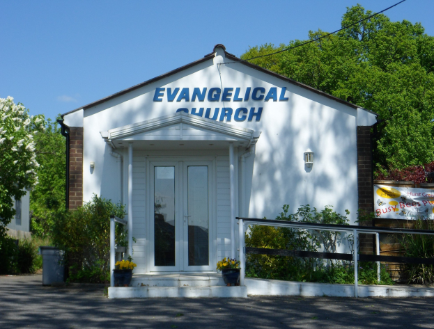 Evangelische Kirche mit Straße davor, umgeben von Grün unter einem klaren blauen Himmel.