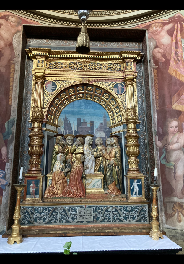 Ein Altar in einer Kirche mit Skulpturen, Kerzen und anderen Gegenständen, mit einem Gemälde an der Wand dahinter und einer Pflanze am unteren Bildrand.