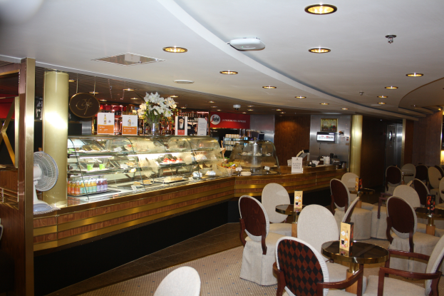 Ein Restaurant-Interieur mit Tischen und Stühlen, einer Theke mit Blumenvasen, Tafeln mit Text, einem Ventilator und anderen Gegenständen unter einer von Lampen beleuchteten Decke.