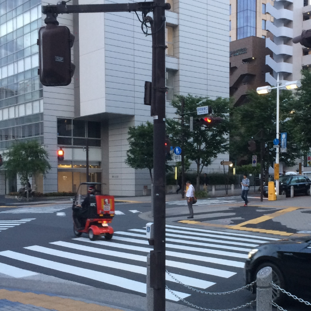 Ein Roller befindet sich auf der Straße mit einigen Personen im Hintergrund, eine Ampel im Vordergrund und Gebäude mit Bäumen im Hintergrund.