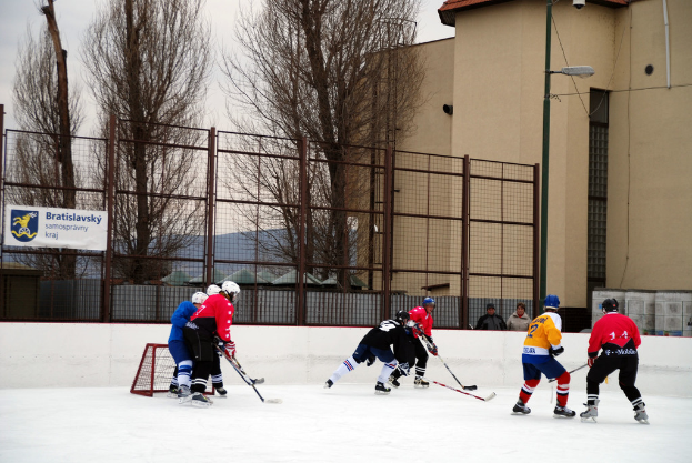 Menschen, die Eis-hockey auf einer Eisfläche mit Gebäuden, Bäumen, einer Straßenlaterne, einer Namensplakette und Zäunen im Hintergrund unter dem Himmel spielen.