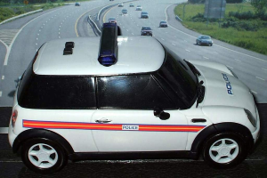 Ein weißer Polizeiwagen ist im Vordergrund geparkt, hinter ihm fahren mehrere Autos auf der Straße, umgeben von Gras und Bäumen auf beiden Seiten, in einer ländlichen Umgebung.