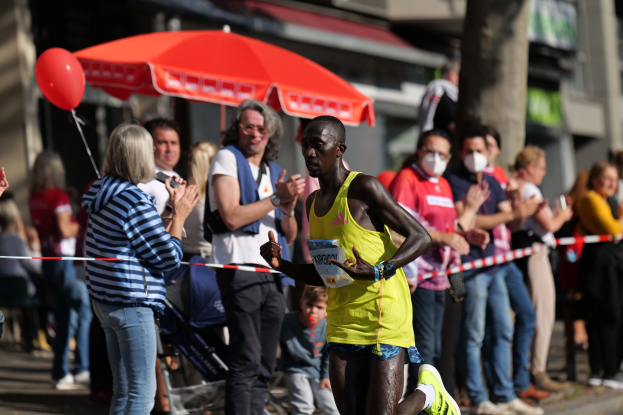Ein Mann in einem gelben T-Shirt und Laufschuhen sprintet durch einen Marathon, während ihn eine Menge umringt und klatscht, mit einem Schirm und einem Kinderwagen im Hintergrund.
