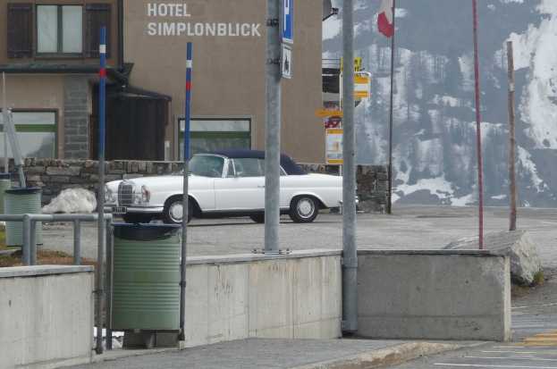 Eine Wand mit Pfählen im Vordergrund, ein Auto und ein Gebäude namens 'Hotel Simplon Blick' in der Mitte und ein schneebedeckter Hügel mit Bäumen im Hintergrund.